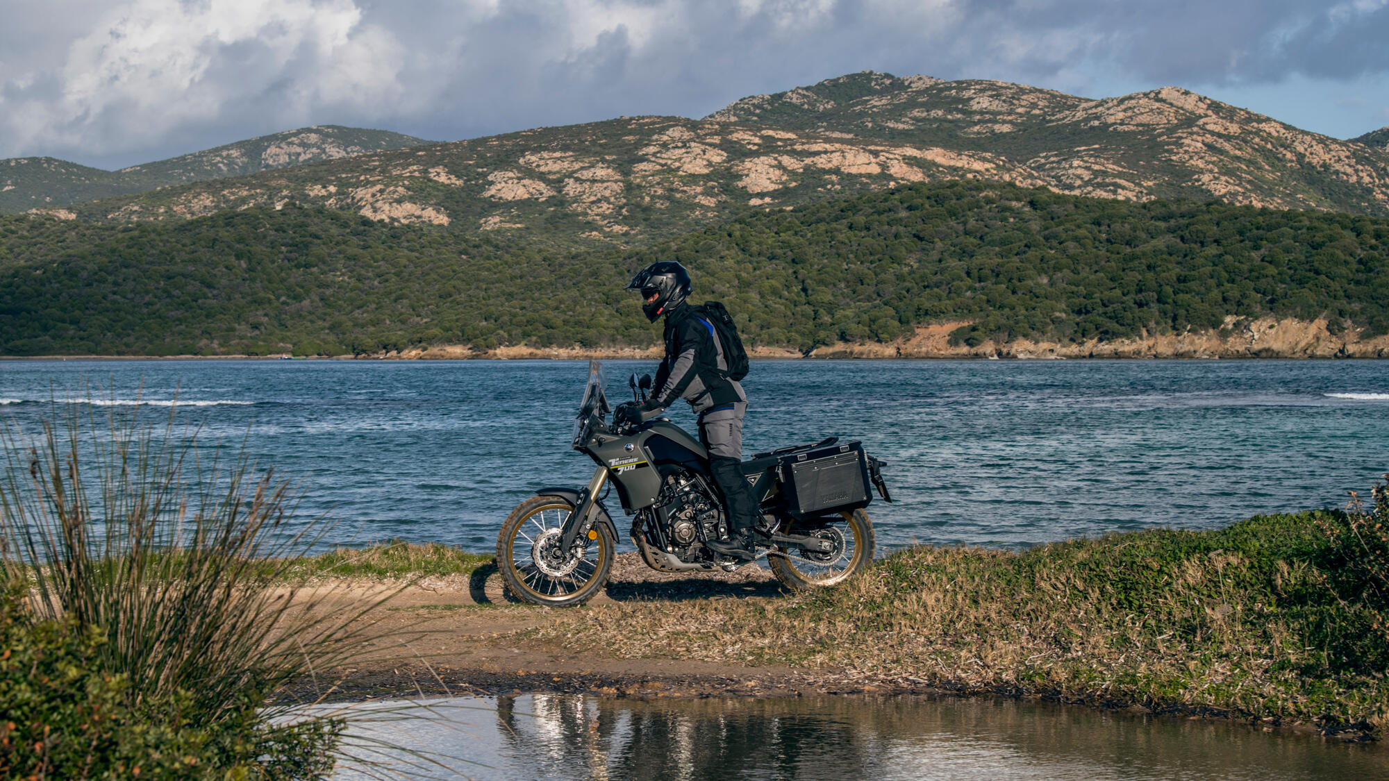A rider rests on the Yamaha Ténéré 700 Explore beside a coastal inlet with side cases fitted and mountains in the distance