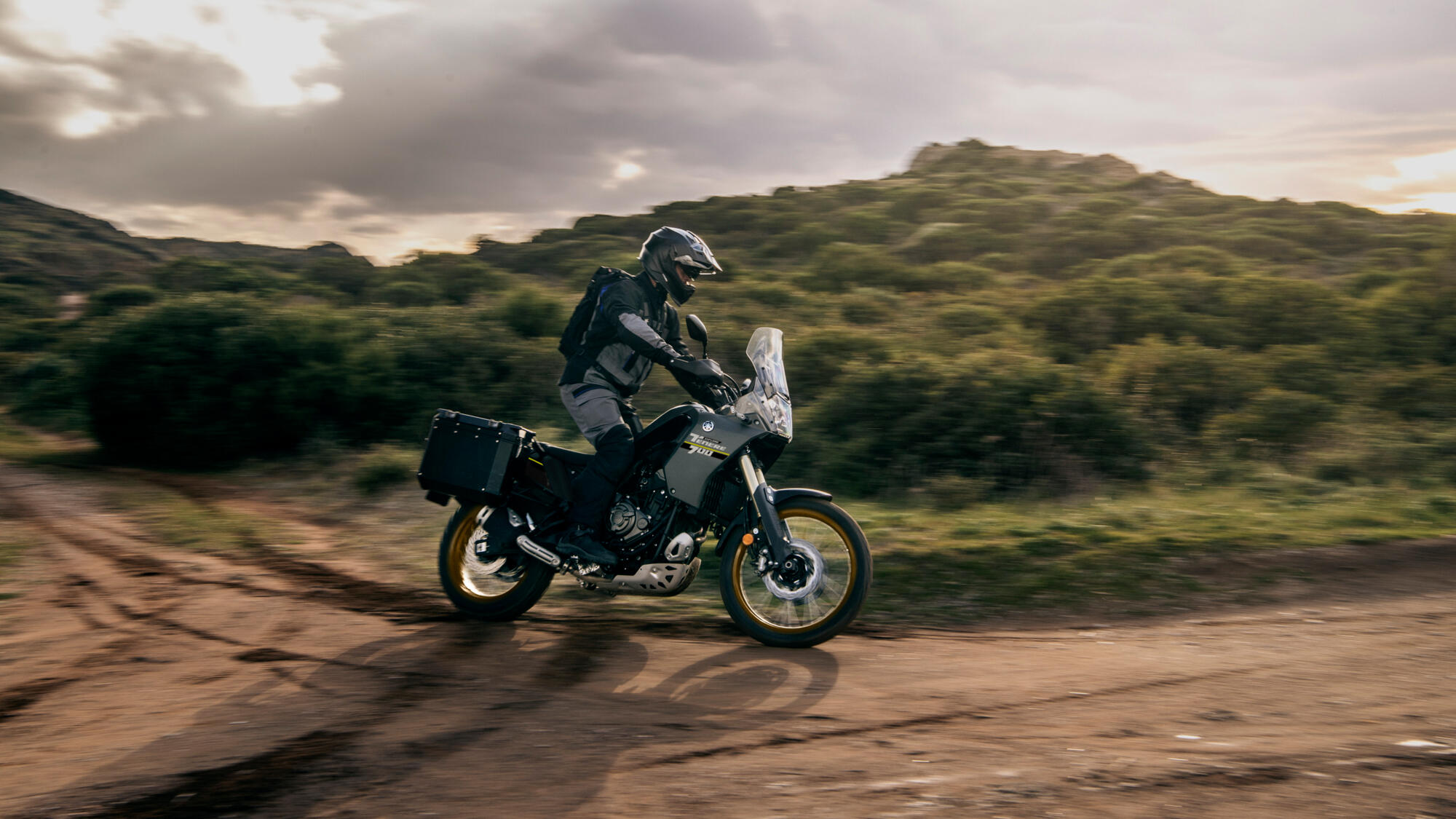 A rider takes the Yamaha Ténéré 700 Explore down a dirt road at dusk with side cases mounted